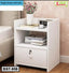 White bedside table with single locking drawer, open shelf, books, flowers, and digital clock.