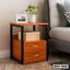 Modern wooden bedside table with black frame, two drawers, and decor items beside a bed