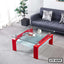 Modern glass center table with red legs in a living room, beside a grey sofa and plant.