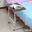 Adjustable laptop table with wheels beside a bed, holding books and a mug