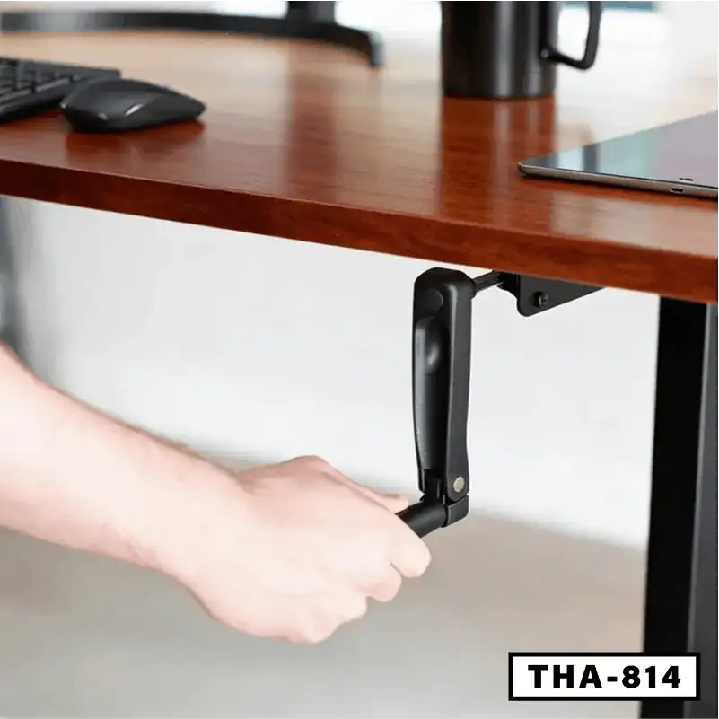 Hand adjusting a wooden height adjustable desk with manual crank handle, office setup visible