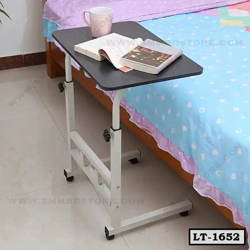 Adjustable laptop table with wheels beside a bed, holding books and a mug Main image